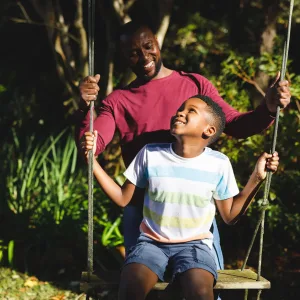 African american father with son having fun and playing on swing in garden. family spending time at home.